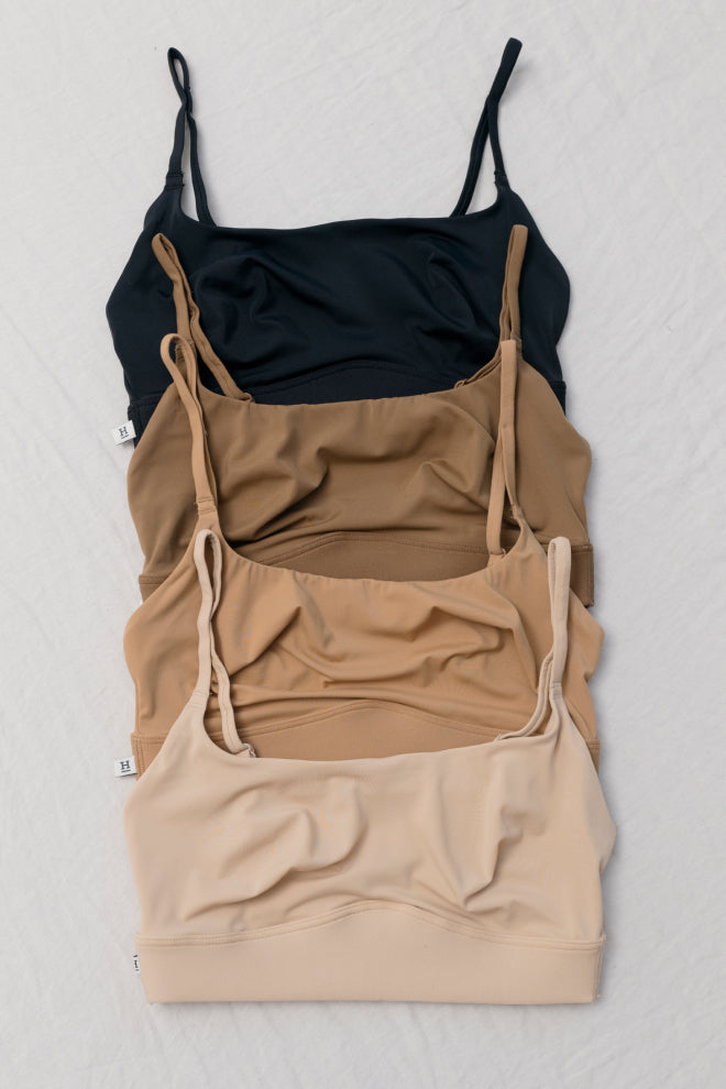 A vertical arrangement of four Bliss Scoop Bralettes in neutral tones, laid out on a flat surface. The bralettes are displayed from top to bottom in Black, Brown, Tan, and Beige, showcasing the variety of colors available in this comfortable and supportive style.