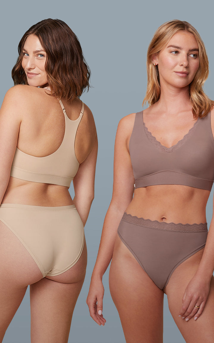 Three models posed against a blue-gray backdrop. Anna stands on the left, facing away to showcase the lace-back detail of the Bliss Lace Halterneck Bralette paired with the Bliss Thong in Black. In the center, Megan wears the Bliss Lace Bralette and Bliss Lace Highwaist Thong in Mauve, facing forward with hands at her sides. On the right, Carli shows the back of the Bliss Racerback Bralette and Bliss Bikini in Beige.