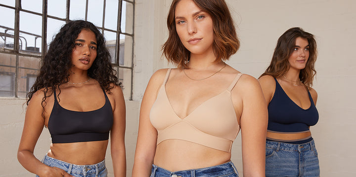 Naisha, Carli, and Summerlyn stand confidently in a bright room with large windows. Naisha wears the Bliss Scoop Bralette in Black with jeans, Carli is in the Bliss Triangle Bralette in Beige with jeans, and Summerlyn wears the Lounge Rib Triangle Bralette in True Blue with jeans. The image highlights the stylish and comfortable fit of the bralettes.