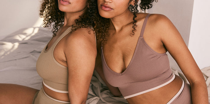 wo models sit closely together on a blanket in a softly lit room. The model on the left, Jade, wears the Lounge Rib Scoop Bralette and Lounge Rib Boyshort in Agave, while the model on the right, Zoe, wears the Lounge Rib Triangle Bralette and Lounge Rib Boyshort in Oyster. Both models exude a relaxed and comfortable vibe, highlighting the cozy and stylish design of the ribbed bralette sets.