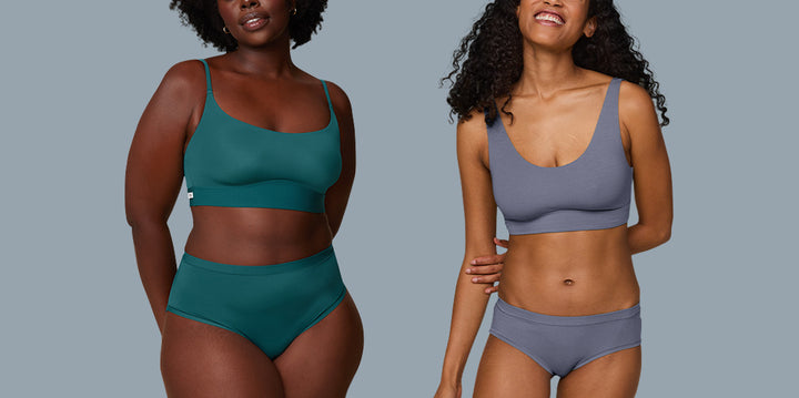 On the left, Akesha wears the Bliss Scoop Bralette and Highwaist Brief in Everglade, standing confidently with a gentle smile. On the right, Zoe models the Cloud Cotton Scoop Bralette and Hiphugger in Slate, smiling brightly with a relaxed posture. Both stand against a soft blue background, highlighting the comfortable and supportive fit of their sets.